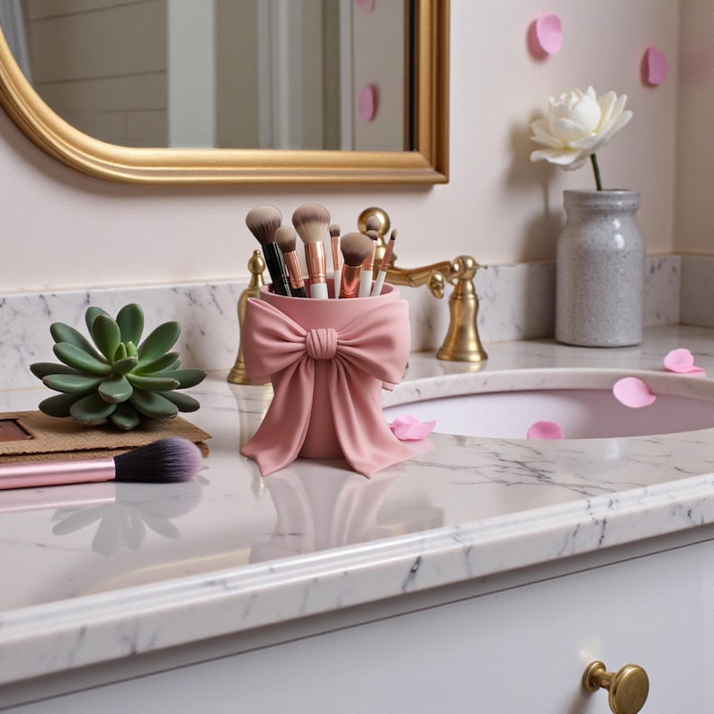 May include: A pink makeup brush holder shaped like a bow, filled with various makeup brushes. The holder sits on a marble countertop next to a succulent and a makeup brush. A gold-framed mirror and faucet are in the background.