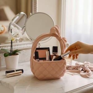 May include: A light pink, quilted-texture storage basket with a prominent bow on the handle. The basket contains various makeup items, including a compact and several tubes. The design features a rounded shape and a structured, solid construction.