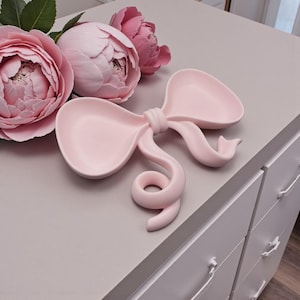 Peut inclure: Un vide-poche en céramique rose pâle en forme de nœud décoratif avec deux larges boucles arrondies et un nœud central. L'objet présente une finition mate et lisse, placé à côté d'un bouquet de trois grandes pivoines rose tendre.