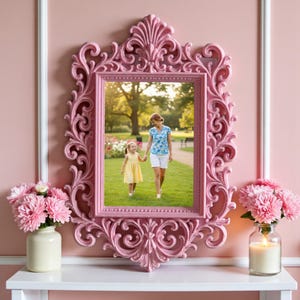 May include: A pink, ornate picture frame with a detailed, scrollwork design. The frame holds a photo of a woman and child walking in a park. The frame is displayed on a white shelf with pink flowers and a candle.