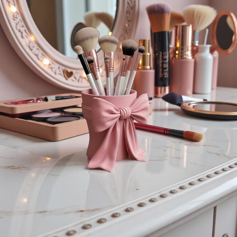 May include: A pink makeup brush holder shaped like a cylinder with a large bow detail. The holder is filled with various makeup brushes. Other makeup items, including a mirror and compact, are visible on the white vanity.