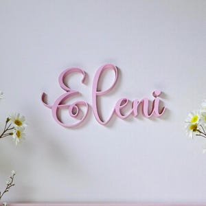 May include: A pink wooden sign in cursive script spelling the name "Eleni". The sign is mounted on a white wall, with a pink shelf below. White flowers with yellow centers are visible on either side of the sign.