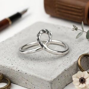 Könnte beinhalten: Ein silberner Ring mit gehämmertem Muster, in Form einer durchgehenden Schleife. Der Ring liegt auf einer grauen Betonoberfläche, im Hintergrund ein Stift, ein ledergebundenes Notizbuch und weitere Ringe. Eine weiße Blume und ein Zweig mit Grünzeug sind ebenfalls zu sehen.
