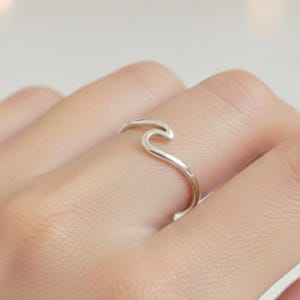 Könnte beinhalten: Ein silberner Ring in Form einer Welle. Der Ring wird an einem Finger getragen, und das Wellendesign ist schlicht und elegant. Der Ring besteht aus glänzendem Metall.