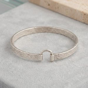 May include: A silver hammered-texture bracelet with a hook clasp. The bracelet is circular and has a textured surface. The clasp is a small, rounded hook that connects the two ends of the bracelet. The bracelet is made of a shiny, metallic material.