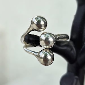 Puede incluir: Un anillo plateado con tres adornos esféricos. El anillo se envuelve alrededor del dedo con las esferas en la parte superior. El anillo se muestra sobre una superficie reflectante.