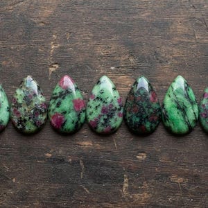 May include: Seven teardrop-shaped cabochons of zoisite with ruby inclusions, arranged in a horizontal row. Each stone features a polished, smooth surface displaying a mottled pattern of vibrant green zoisite, black hornblende, and deep red ruby.