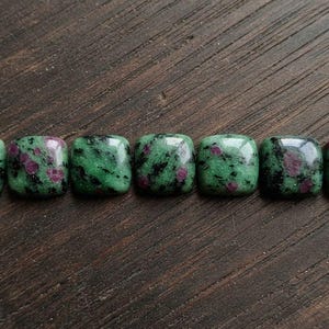 Peut inclure: Sept cabochons carrés en zoïsite avec des inclusions de rubis, présentant une surface polie avec un motif tacheté de vert vif, de noir profond et de rose magenta, disposés en une rangée horizontale sur une surface en bois sombre.