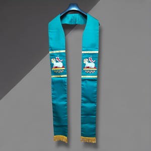 May include: Teal graduation stole with gold trim and a detailed embroidered design featuring a lamb and a cross. The stole has a gold fringe at the bottom and is hanging on a blue hanger.