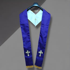 Puede incluir: Una estola púrpura con flecos dorados y cruces bordadas. La estola está drapeada sobre un cuello blanco con una cruz dorada. La estola está hecha de una tela brillante y está colgada de una percha negra.