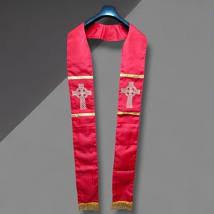 May include: A red clergy stole with white embroidered Celtic crosses and gold trim. The stole hangs from a black hanger against a gray background. The bottom of the stole has gold fringe.