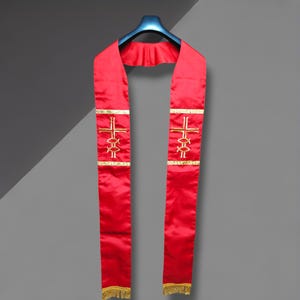 May include: A vibrant red clergy stole with gold embroidered crosses and fish symbols. The stole has a gold trim and a gold fringe at the bottom. The stole is hanging on a black hanger against a gray background.