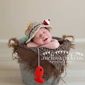 May include: A newborn baby wearing a crocheted fishing hat with a red and white bobber on top, sleeping in a metal bucket lined with burlap. A red crocheted fish is hanging from a string.