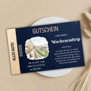 Op de afbeelding: Een marineblauwe cadeaubon met gouden tekst en accenten. De bon luidt "GUTSCHEIN" en "FÜR EINEN Wochenendtrip". Het bevat een aquarel illustratie van een Europese stad en de tekst "ALLES GUTE!"