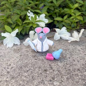 Könnte beinhalten: Eine graue Mäusefigur mit rosa Ohren und einem weißen Laborkittel, die eine blau-weiße Spritze hält. Daneben befinden sich ein rosa Block und eine blaue Flasche. Die Szene spielt auf einer Steinoberfläche mit weißen Blumen und grünem Laub.