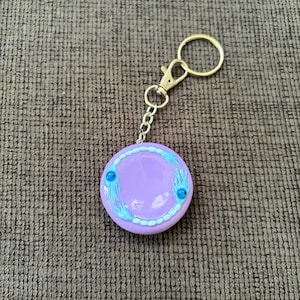 May include: A circular lavender keychain featuring a metallic silver split ring and lobster clasp. The centre displays a light blue beaded border with two small, blue, tentacled creatures positioned on opposite sides of the smooth, glossy surface.