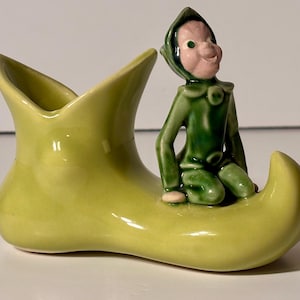 May include: A ceramic figurine of a seated elf in a green outfit with a pink face, perched on a light green shoe-shaped planter. The planter has a flared opening.
