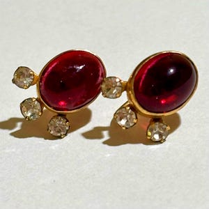 May include: A pair of gold-tone stud earrings featuring a large, smooth, oval-shaped deep red cabochon stone. Each earring is accented with three small, clear, round-cut faceted stones arranged in a cluster along the side of the red center piece.