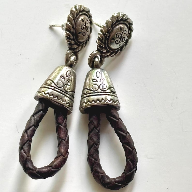 Op de afbeelding: Een paar hangende oorbellen met een cirkelvormige zilverkleurige knop met een gedraaide touwrand en een gegraveerd midden. Een zilverkleurig klokvormig onderdeel met gegraveerde patronen is verbonden met een donkerbruine gevlochten lederen lus.