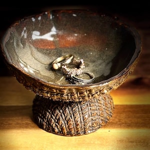 May include: A handcrafted ceramic jewelry dish with a textured, brown base and a bowl with a speckled, earthy glaze. Two rings rest inside the bowl. The dish is designed to hold rings and small jewelry items.