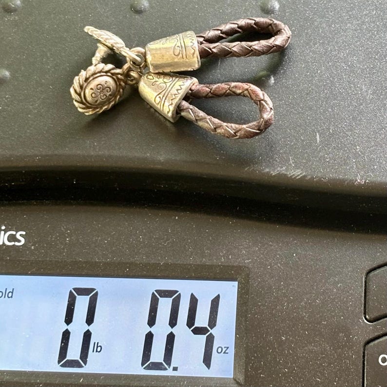 Op de afbeelding: Een paar oorbellen met donkerbruine gevlochten leren lussen bevestigd aan zilverkleurige metalen kapjes met gegraveerde patronen en ronde knoppen. De oorbellen liggen op een digitale weegschaal die 11,3 gram aangeeft.