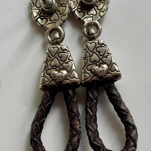 Op de afbeelding: Een paar hangende oorbellen met een zilverkleurige metalen knop met een hartjespatroon. Daaronder hangt een bijpassend driehoekig zilverkleurig metalen stuk met een hartmotief, verbonden met een donkerbruine gevlochten leren lus aan de onderkant.