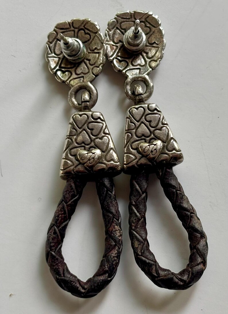 Op de afbeelding: Een paar hangende oorbellen met een zilverkleurige metalen knop met een hartjespatroon. Daaronder hangt een bijpassend driehoekig zilverkleurig metalen stuk met een hartmotief, verbonden met een donkerbruine gevlochten leren lus aan de onderkant.