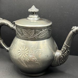 May include: A silver-toned metal teapot featuring an engraved floral pattern on the body, a lid with a decorative finial, and an ornate handle and spout. The upper section of the teapot is adorned with a relief band of grape leaves and vines.