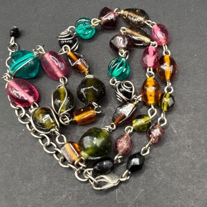 May include: A multi-strand necklace featuring a silver-toned metal chain interspersed with various glass beads in shades of emerald green, amber, magenta, and black. Some beads are wrapped in thin silver wire, and the necklace includes a lobster clasp.