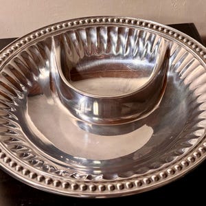May include: A silver-colored serving platter with a central dip bowl. The platter has a beaded edge and a fluted design around the dip bowl. The surface is reflective, and the overall shape is round.