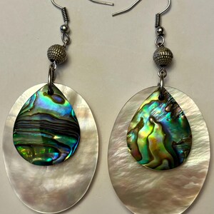 May include: A pair of dangle earrings featuring iridescent abalone shell teardrop shapes set on oval mother-of-pearl backgrounds. Each earring has a silver-tone metal hook and a textured bead accent.