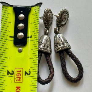 Op de afbeelding: Een paar zilverkleurige metalen oorbellen met een ronde knop met etswerk, een klokvormige hanger en een lus van donkerbruin gevlochten leer. Een geel meetlint ligt ernaast en geeft een lengte van 5 cm aan.