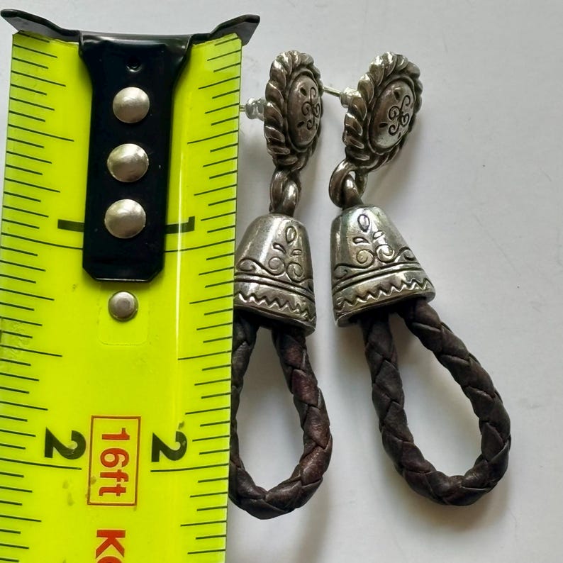 Op de afbeelding: Een paar zilverkleurige metalen oorbellen met een ronde knop met etswerk, een klokvormige hanger en een lus van donkerbruin gevlochten leer. Een geel meetlint ligt ernaast en geeft een lengte van 5 cm aan.