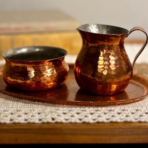 May include: A copper pitcher and bowl set with hammered texture. The pitcher has a handle and a silver-colored rim. The bowl is round with a similar hammered texture and silver-colored rim. Both pieces sit on a copper tray.