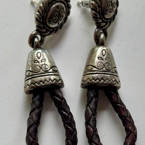 Op de afbeelding: Een paar oorbellen met een zilverkleurige metalen knop met een gedraaide rand en een gegraveerd midden. Een klokvormige zilverkleurige metalen kap met bloem- en zigzagpatronen is verbonden met een donkerbruine gevlochten leren lus.