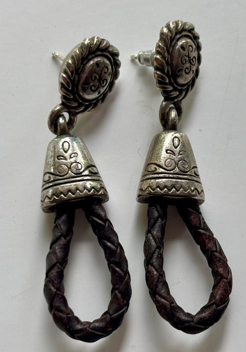 Op de afbeelding: Een paar oorbellen met een zilverkleurige metalen knop met een gedraaide rand en een gegraveerd midden. Een klokvormige zilverkleurige metalen kap met bloem- en zigzagpatronen is verbonden met een donkerbruine gevlochten leren lus.