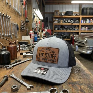 May include: A gray and black trucker hat with a leather patch that reads "THAT'S WHAT I DO, I FIX STUFF AND I KNOW THINGS." The hat is sitting on a wooden workbench with tools.