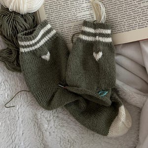 Könnte beinhalten: Ein offenes Buch liegt auf einer weichen, weißen Decke neben einem Paar olivgrüner Stricksocken mit weißen Streifen und Herzdetails. Garn in Weiß und Olivgrün ist ebenfalls vorhanden.