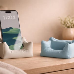Könnte beinhalten: Zwei Telefonständer in weichem, kissenähnlichem Design. Einer ist cremefarben und hält ein Telefon mit einem grünen und blauen Landschaftshintergrund. Der andere Ständer ist hellblau. Beide stehen auf einer Holzoberfläche.