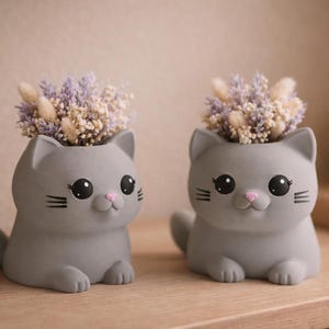 Könnte beinhalten: Zwei graue, katzenförmige Pflanzgefäße mit großen schwarzen Augen, rosa Nasen und kleinen schwarzen Schnurrhaaren. Jedes Pflanzgefäß enthält einen Strauß getrockneter Blumen in Lila, Beige und Weiß. Auf einer Holzoberfläche.