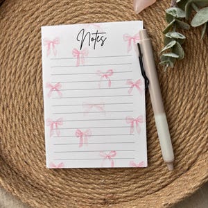 Könnte beinhalten: Ein Notizblock mit rosa Schleifenmotiven und dem Wort "Notes" in Schreibschrift. Die linierten Seiten sind mit Aquarellschleifen verziert. Ein beiger Stift mit schwarzem Clip liegt neben dem Notizblock. Der Notizblock liegt auf einem geflochtenen Tablett.