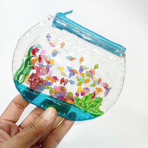 Puede incluir: Un monedero transparente con forma de pecera con cremallera y ribete azules. El monedero está lleno de confeti colorido con forma de pez y corales y algas bordadas. El diseño es caprichoso y juguetón.