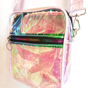Könnte beinhalten: Schultertasche aus schimmerndem, transparentem Material mit rosa Besatz und Riemen. Die Tasche hat eine Reißverschluss-Vordertasche mit einem Regenbogen-Reißverschluss. Der Riemen ist hellrosa mit einem Herzmuster. Die Tasche ist zum Tragen über der Schulter konzipiert.