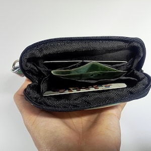Peut inclure: Un petit portefeuille noir zipp&eacute; est ouvert, r&eacute;v&eacute;lant plusieurs compartiments. &Agrave; l'int&eacute;rieur, il y a plusieurs cartes et un billet vert. Le portefeuille a un petit anneau argent&eacute; attach&eacute; sur le c&ocirc;t&eacute;.