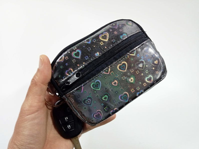 Peut inclure: Un petit porte-monnaie rectangulaire avec une fermeture &eacute;clair noire et une finition holographique argent&eacute;e. Le sac est orn&eacute; d'un motif de c&oelig;urs color&eacute;s. Une cl&eacute; de voiture est attach&eacute;e au sac.