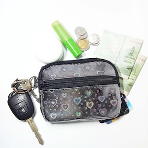 Peut inclure: Une petite pochette noire zipp&eacute;e avec un motif de c&oelig;urs, des cl&eacute;s, des pi&egrave;ces de monnaie, des billets de banque et un tube vert. La pochette est ouverte, r&eacute;v&eacute;lant des cartes &agrave; l'int&eacute;rieur. Les cl&eacute;s sont attach&eacute;es &agrave; un porte-cl&eacute;s.