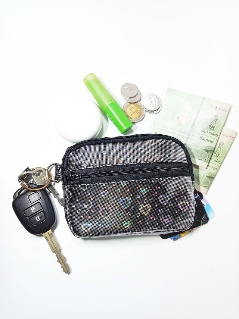 Peut inclure: Une petite pochette noire zipp&eacute;e avec un motif de c&oelig;urs, des cl&eacute;s, des pi&egrave;ces de monnaie, des billets de banque et un tube vert. La pochette est ouverte, r&eacute;v&eacute;lant des cartes &agrave; l'int&eacute;rieur. Les cl&eacute;s sont attach&eacute;es &agrave; un porte-cl&eacute;s.