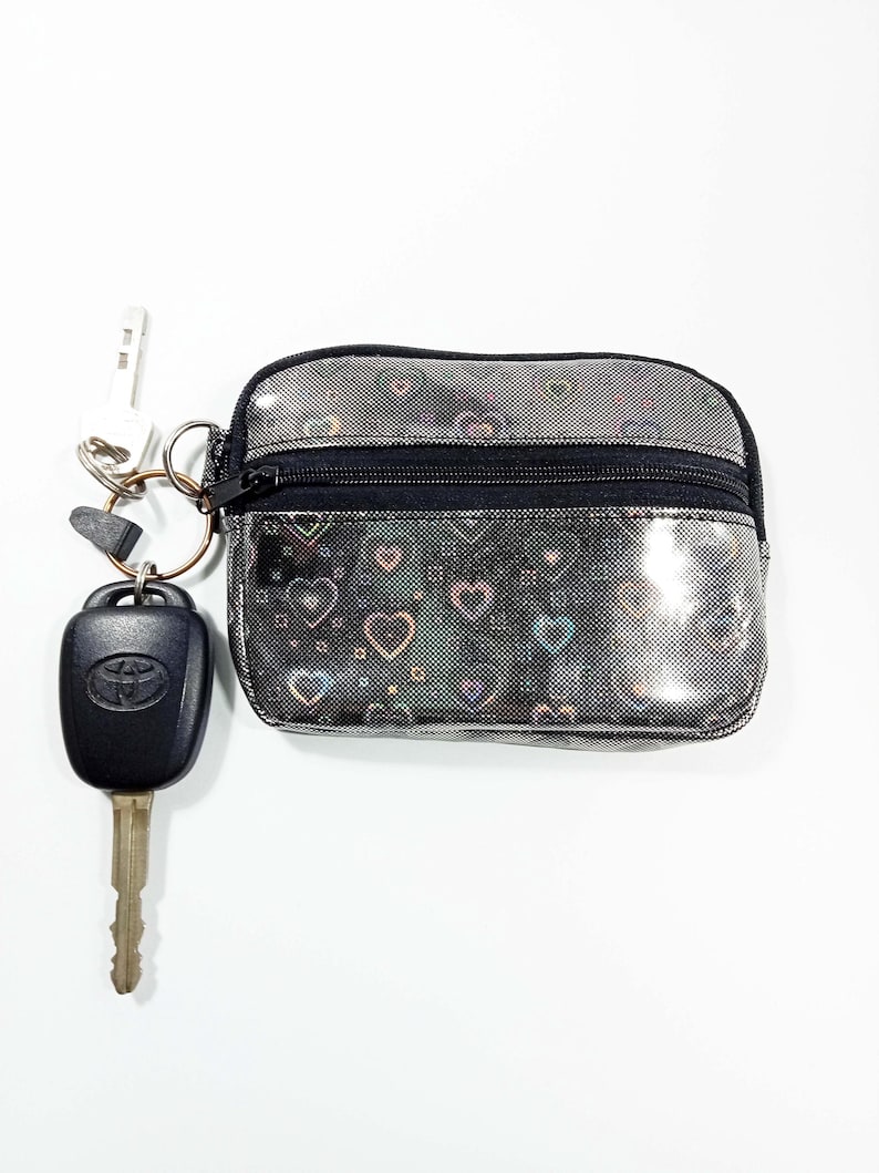 Peut inclure: Petite pochette argent&eacute;e avec fermeture &eacute;clair noire et motif c&oelig;urs. Un porte-cl&eacute;s avec une cl&eacute; de voiture et une cl&eacute; de maison est attach&eacute;. La pochette est con&ccedil;ue pour contenir de petits objets comme des cl&eacute;s, des cartes ou de l'argent.