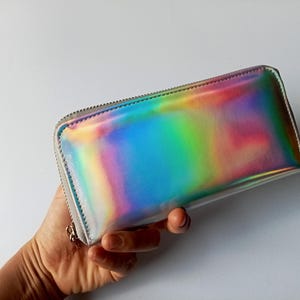 Könnte beinhalten: Eine rechteckige Geldbörse mit regenbogenfarbenem, schillerndem Finish. Die Geldbörse hat einen silbernen Reißverschluss und einen silbernen Zipper. Die Geldbörse wird in einer Hand gehalten.