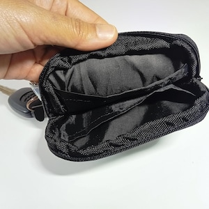 Peut inclure: Petite pochette &agrave; cl&eacute;s noire zipp&eacute;e ouverte, avec des poches int&eacute;rieures. Un jeu de cl&eacute;s est visible &agrave; c&ocirc;t&eacute; de la pochette ouverte. La pochette est en tissu textur&eacute; et r&eacute;sistant.